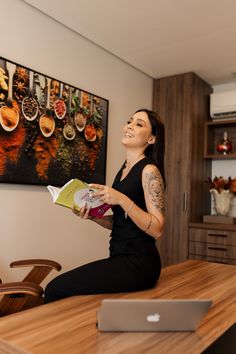 a woman sitting at a table with a laptop and book in her hand, reading a cookbook