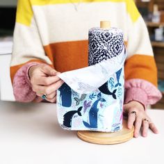 a woman is wrapping fabric around a bottle on a wooden stand while another person sits at the table behind her