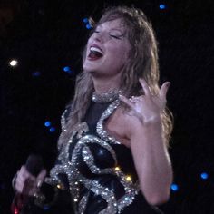 a woman singing into a microphone with her hands in the air and lights on behind her