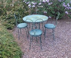 an outdoor table and chairs in gravel area next to bushes with purple flowers behind them