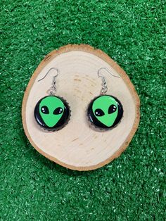 two green alien earrings sitting on top of a piece of wood with grass in the background