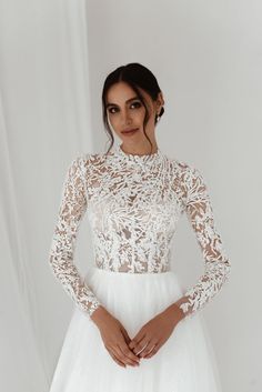 a woman wearing a white wedding dress with sheer lace on the neck and sleeves, standing in front of a white backdrop