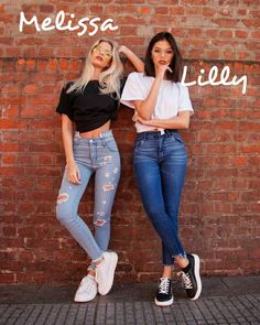 two women standing next to each other in front of a brick wall wearing ripped jeans