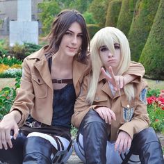 two people sitting on a bench in front of some flowers and bushes with one pointing at the camera