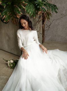 a woman in a white dress sitting on the ground next to a plant and flowers