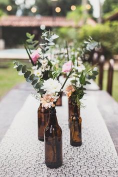 three brown bottles with flowers in them sitting on a long white tableclothed runner