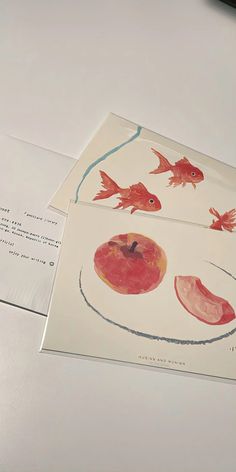 two cards with fish and apples on them are sitting on a table next to an envelope