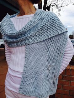 a woman wearing a blue knitted shawl standing next to a brick wall in front of a tree