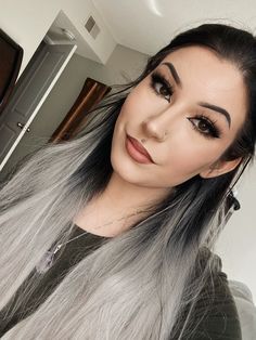 a woman with long grey hair and black eye makeup looks at the camera while standing in front of a mirror