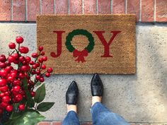 a person standing in front of a door mat with the word joy on it and a christmas wreath