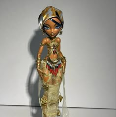 a doll is dressed in gold and has blue eyeliners on her face, holding a purse