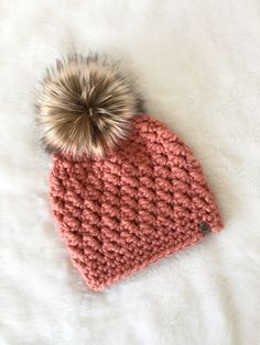 a knitted hat with a pom - pom sits on a white blanket