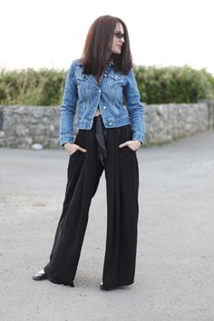 a woman wearing black wide legged pants and a jean jacket is standing in the street