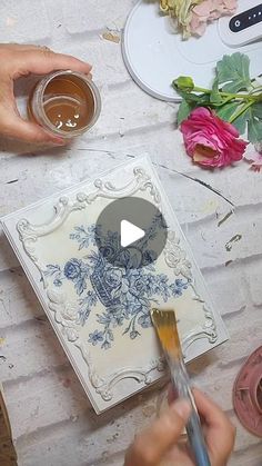 two hands holding paintbrushes over a piece of paper with flowers on the table