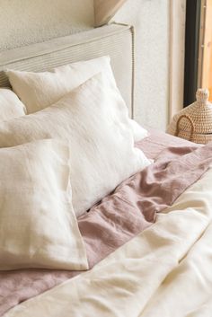 a bed with white and pink linens in front of an open window, next to a wicker basket