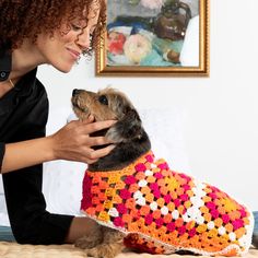 a woman petting a small dog wearing a crocheted sweater on top of a bed