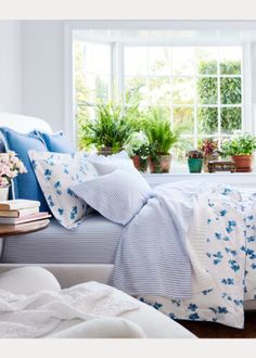 a bed with blue and white comforters in front of a window filled with potted plants