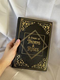 a hand holding a book that is black with gold trimmings and the title, a court of thorns & roses