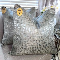 two decorative pillows with tags on them sitting on a couch in front of a window