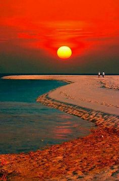 two people are walking on the beach as the sun sets in the sky over the water