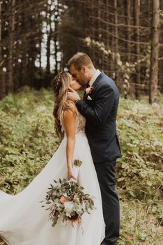 a bride and groom standing in the woods kissing each other with their arms around each other