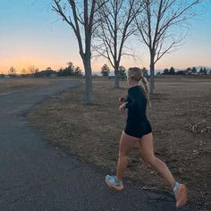 a woman running down a road near some trees and grass with the sun setting in the background