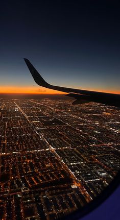 an airplane wing flying over a city at night with the sun setting in the distance