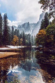 a lake surrounded by trees with mountains in the background and snow on the ground around it