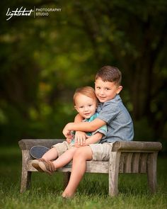 two young boys sitting on a wooden bench in the grass with their arms around each other