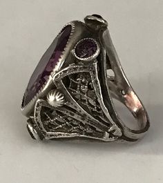 "This lovely filigreed and ornate ring has an oval shaped faceted amethyst purple color glass stone at its center. It is flanked on either side of to and bottom with small round bezel set purple glass stones. The sides have lattice shaped cut outs and the band is smooth. The face of this ring is about 3/4\" long and about 1/2\" across. It is a size 3 1/4 and is marked Sterling. Please note there are some overall signs of wear, especially to the stones, four of five of which, including the center Antique Purple Amethyst Ring With Accent Stones, Oval Amethyst Jewelry Collectible, Antique Oval Purple Jewelry, Antique Purple Oval Jewelry, Ornate Oval Amethyst Ring, Oval Sterling Silver Filigree Amethyst Ring, Victorian Style Oval Purple Amethyst Ring, Oval Amethyst Collectible Ring, Silver Amethyst Ring With Intricate Design