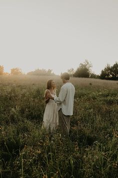 a man and woman standing in the middle of a field at sunset, looking into each other's eyes