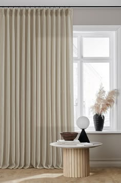 a room with a table, vase and curtain in front of a window that is open