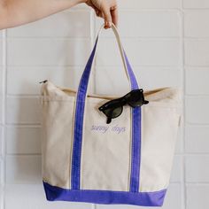 a person holding a white and blue tote bag with sunglasses on the handle, in front of a brick wall