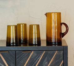 three coffee mugs sitting on top of a wooden cabinet next to a vase and pitcher