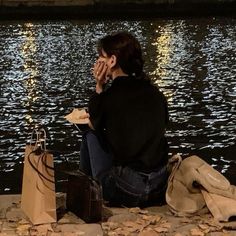 a woman sitting on the ground next to water with her hands in her face and shopping bags