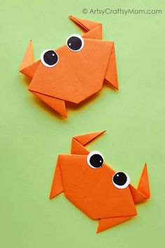 two orange origami fish with googly eyes on green paper and one has black eyes