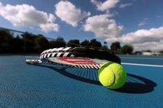 a tennis ball and racket on a blue court with clouds in the sky behind it