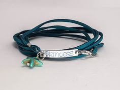 "Hand-stamped wrap bracelet for a favorite princess. Fitted as a 7\" to a 7-1/2\" bracelet, it adjusts smaller or larger depending on how many loops you make. Bracelet is a long strand of teal faux suede--so the color will not bleed--with a stamped aluminum plate with blue lettering. Lobster claw clasp makes it easy to latch. Coordinating necklace is also available. Choose between Pinafore or Fantasy lettering. Not sure how to use it? See a video of how to wear the bracelet at https://youtu.be/B Adjustable Nickel-free Name Bracelet, Handmade Adjustable Green Name Bracelet, Adjustable Nickel Free Name Bracelet, Nickel-free Adjustable Bangle Wrap Bracelet, Adjustable Wrap Bracelet For Friendship, Adjustable Hand Stamped Bracelets, Adjustable Hand Stamped Bracelet, Adjustable Hand Stamped Bohemian Bracelets, Bohemian Adjustable Hand Stamped Bracelets