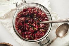 a glass jar filled with cranberry sauce next to two spoons on top of a white cloth