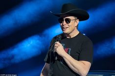 a man wearing a cowboy hat and sunglasses holding a microphone in front of a stage