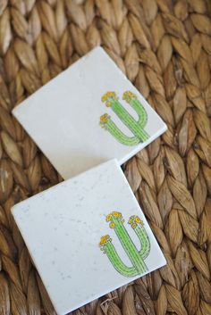 two coasters with cactus designs on them sitting on a woven tablecloth covered surface