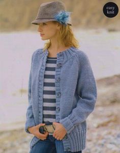 a woman wearing a hat and cardigan standing next to the water with her hands in her pockets