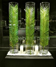 three tall glass vases filled with green leaves and candles sitting on a white surface