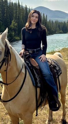 a woman riding on the back of a brown horse next to a lake and forest