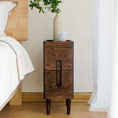 a wooden nightstand with a plant on it next to a bed and white sheets in a bedroom