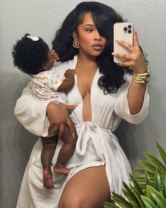 a woman holding a baby and taking a selfie with her cell phone while wearing a white dress