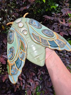 a hand holding a green butterfly shaped object in front of some bushes and trees with purple leaves