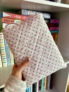 a person holding up a quilted pillow in front of bookshelves