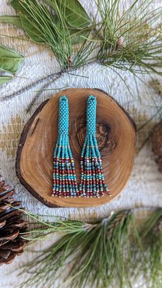 two pairs of beaded earrings sitting on top of a piece of wood next to pine cones