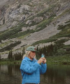 a woman standing in front of a lake taking a photo with her cell phone while wearing a blue jacket and hat